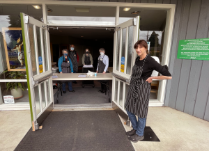 The doors are swung wide open to Maple Alley Inn, with the door held open by a smiling woman in an apron. In the background a group of volunteers in safety masks stands eager and welcoming.