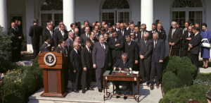 President Lyndon B. Johnson signs a document at a desk in front of many people in black suits in front of the White House.