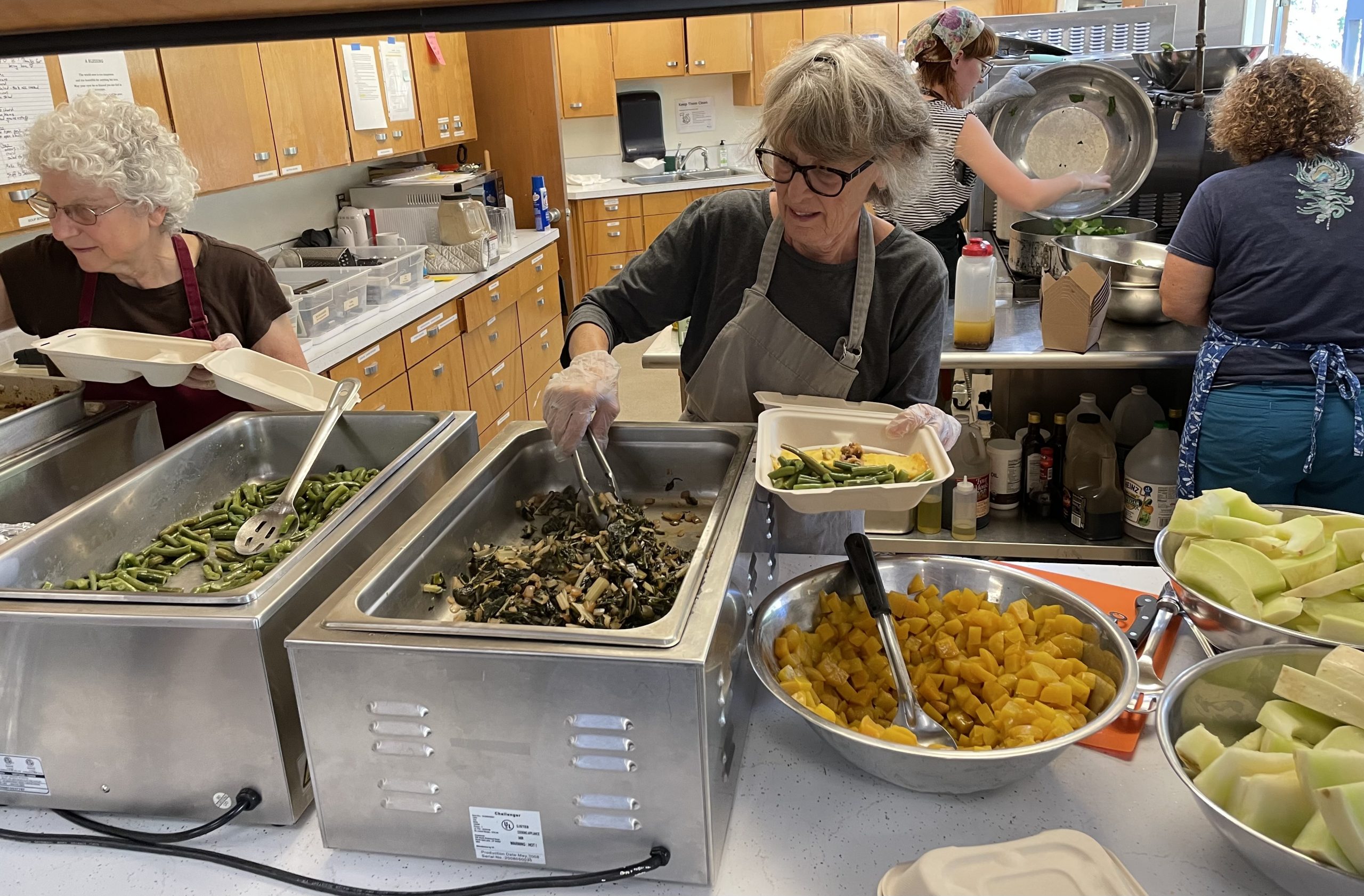Four women in aprons, volunteers at the Maple Alley Inn kitchen, stand in the kitchen preparing and doling out food for guests to the hot meals program.