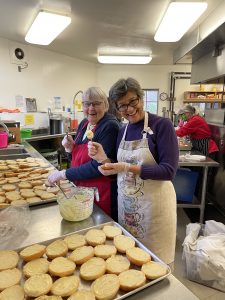 Two women smile together in a kitchen as they spread sauce onto bread buns