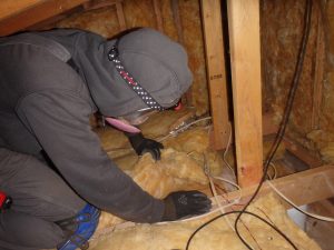 A man wearing goggles crouches in an attic looking at the insulation.