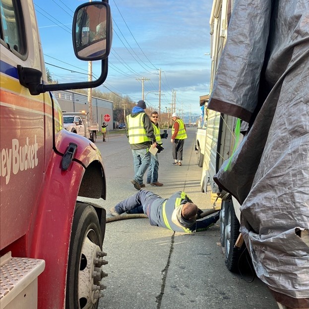A Honey Bucket truck is parked alongside an RV, as people in neon yellow vests stand by and a man wearing neon-striped gear lies in the street, pumping black water with a hose from the bottom of an RV.