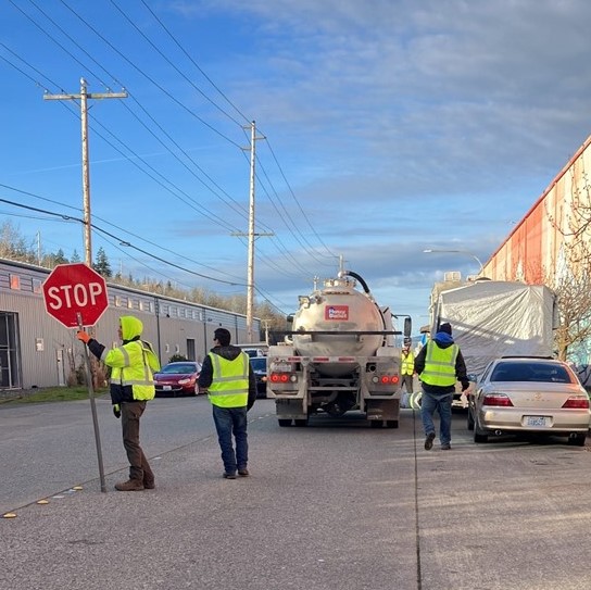 People in neon yellow gear stand in a street, one holding a Stop sign to direct traffic, two walking alongside a large Honey Bucket truck.