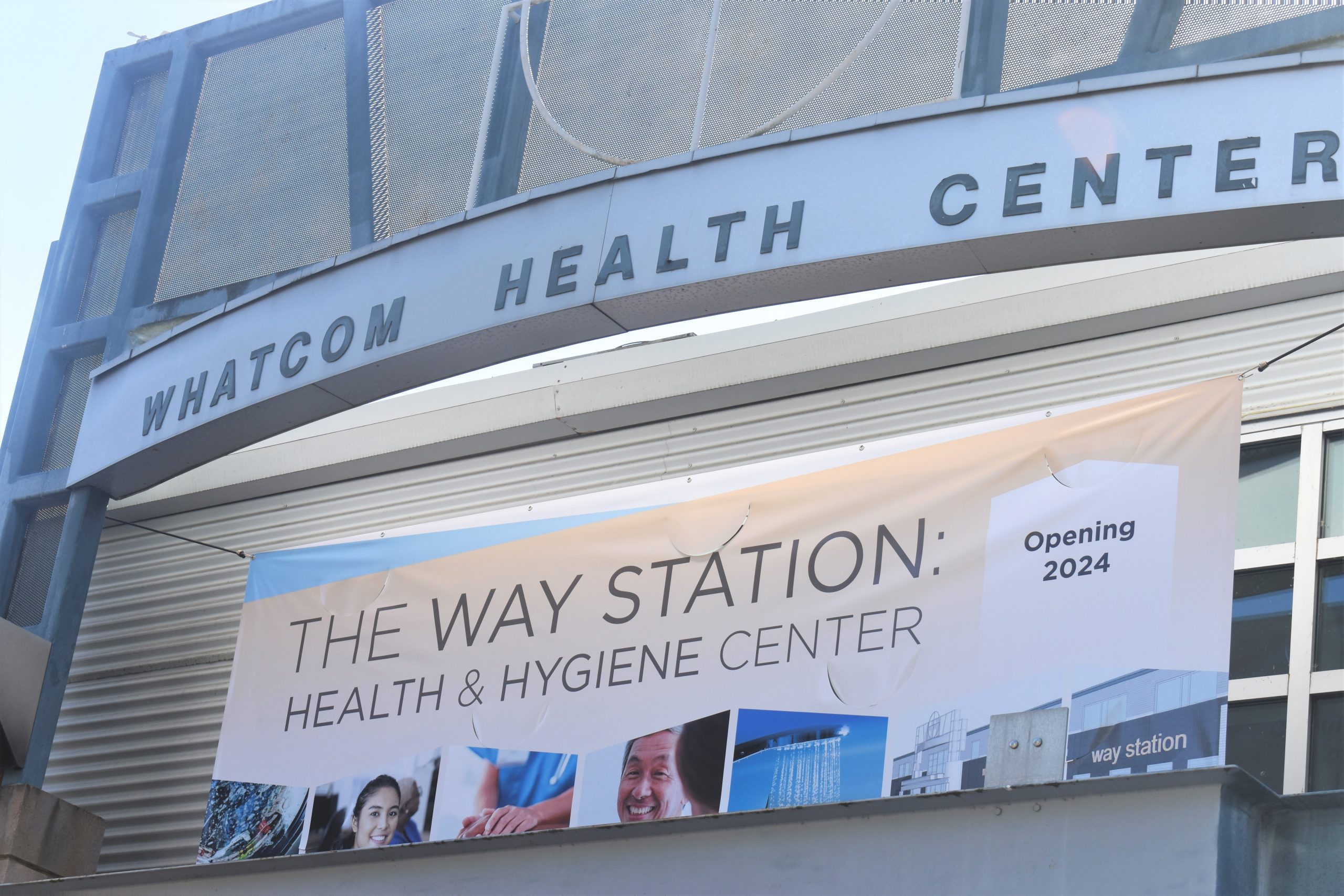 A silver metallic building awning reads "Whatcom Health Center" in all caps. On the front of the building, a vinyl banner says "The Way Station."