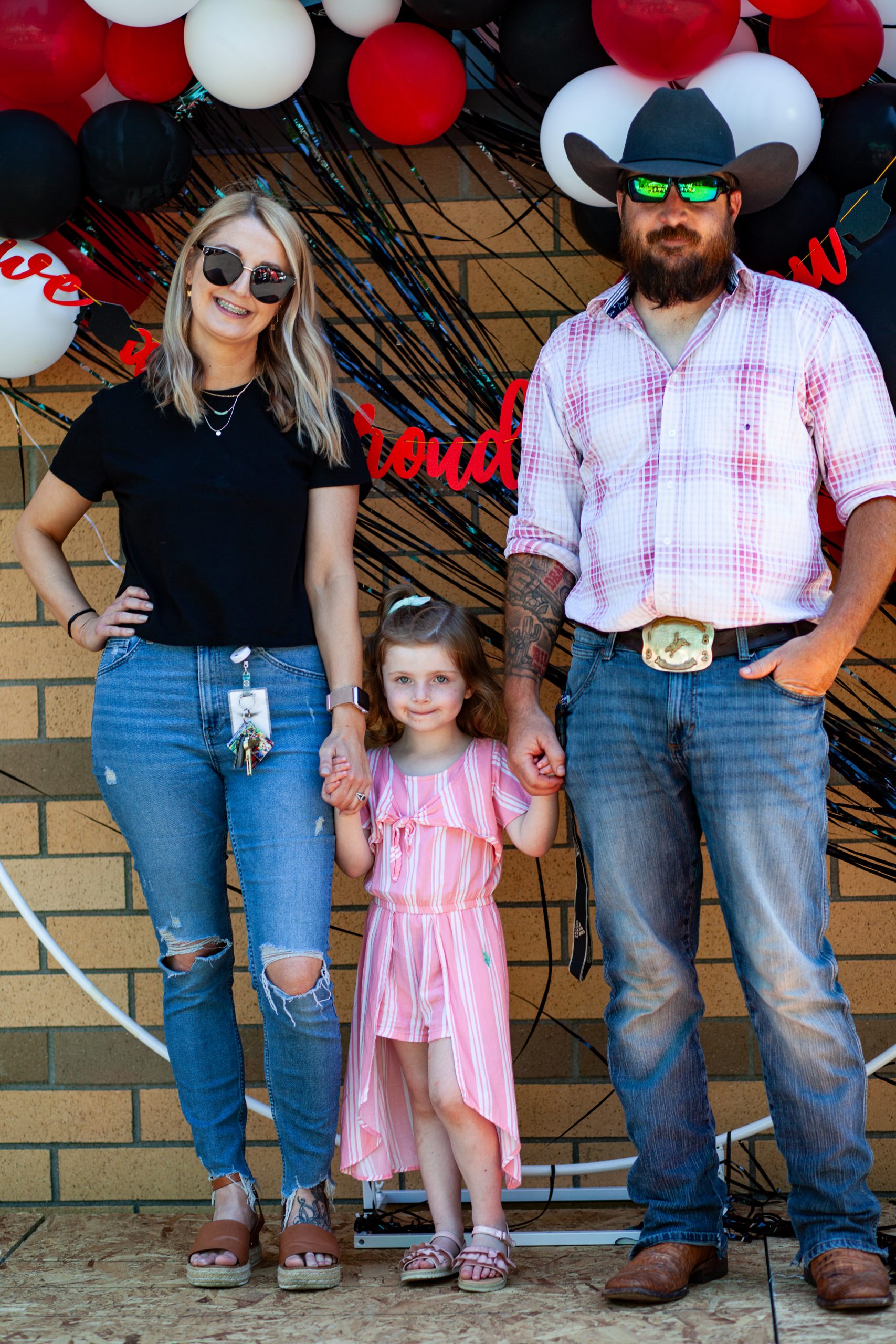 A woman, young girl, and man stand contentedly, the man and woman each holding one of the girl's hands.