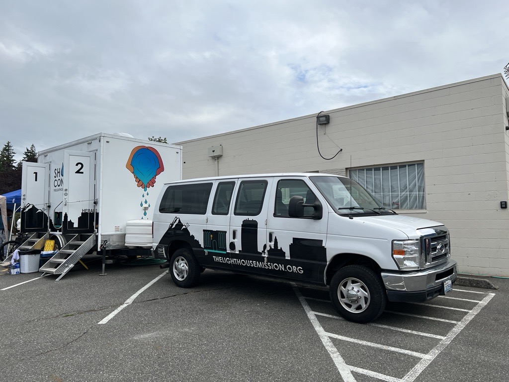 A van with a silhouette graphic and the text thelighthousemission.org is parked in front of a trailer with two doors and two sets of metal stairs.