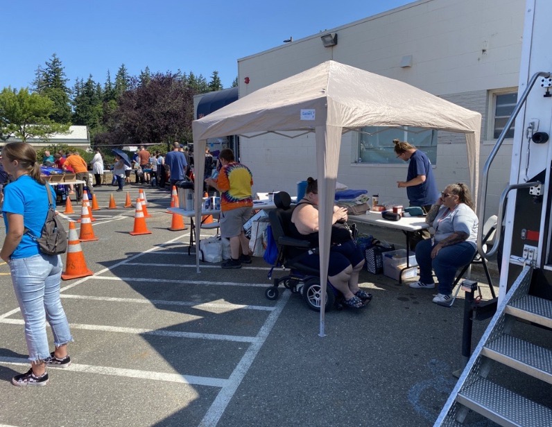 People, who are members of Opportunity Council's Homeless Outreach Team, stand under the shade of a fabric tent around a folding table helping a client in a wheelchair. The corner of a trailer, the Shower Connect Truck, is visible in the frame. Orange construction cones are visible in the background, in the parking lot where the tent and trailer are located, and people gather near the entrance of the warehouse building in the background, the Salvation Army.