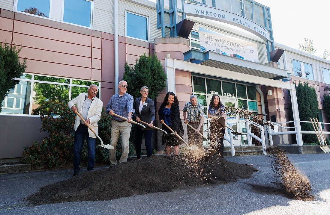 A group of people in professional clothing smile and laugh as they shovel dirt from a long pile in front of a building, which has a banner that says "The Way Station: Health & Hygiene Center" and shows the address 1500 N. State St.