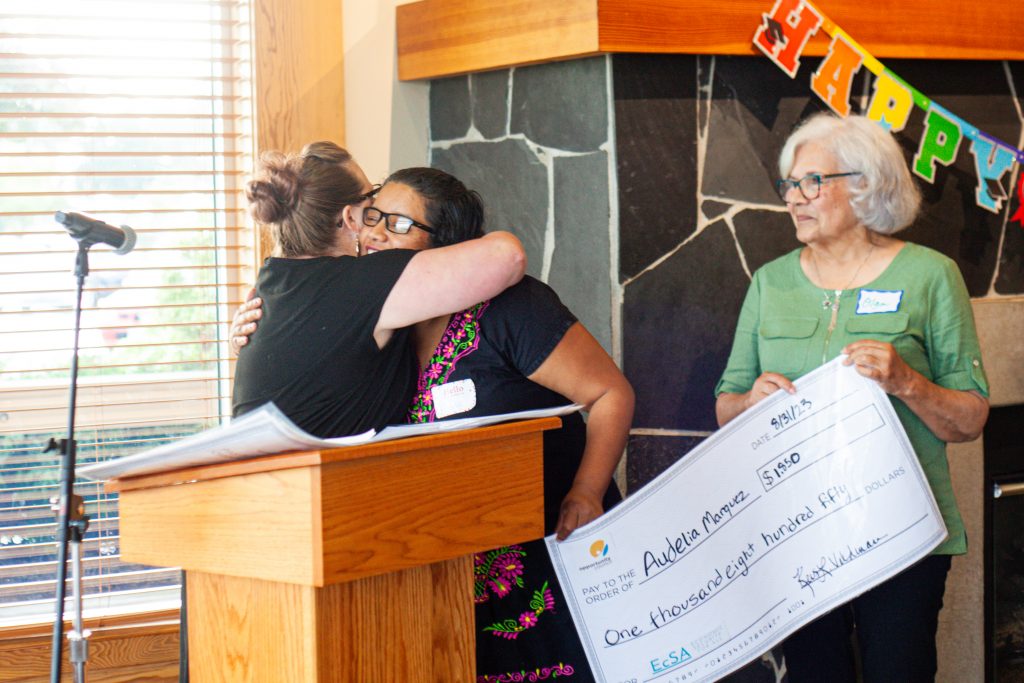 Two women hug at a pulpit and a third stands by looking on with an oversize check that says "8/31/23: $1,850: One thousand eight hundred fifty Dollars". In the "To" line is the EcSA logo with the acronym in teal font. In the top left is the Opportunity Council teal and gold logo.