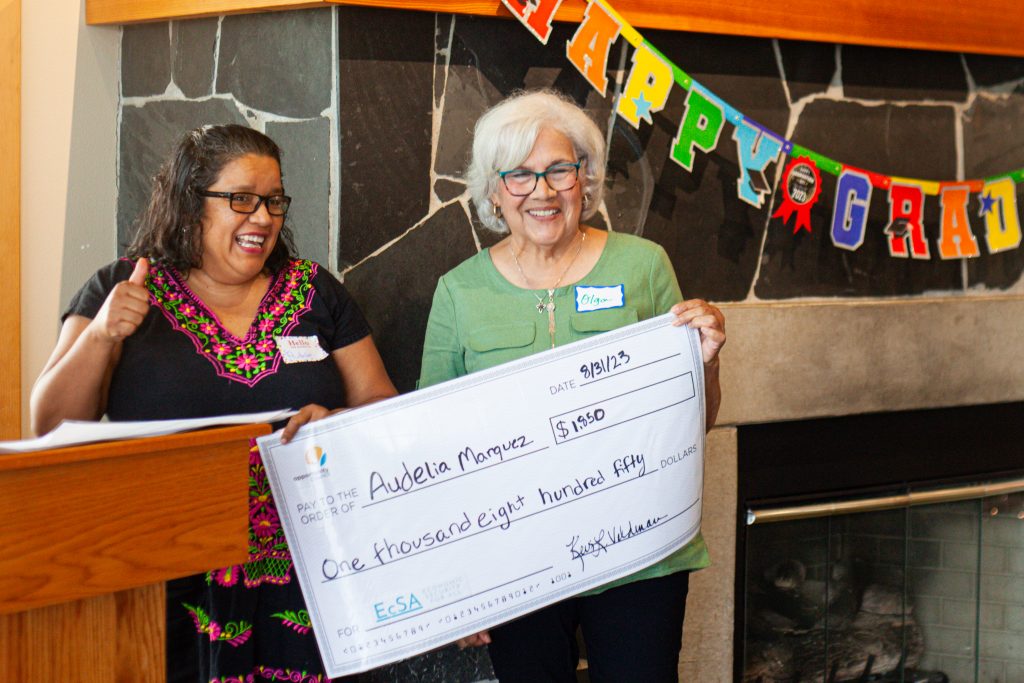 Two women smile elatedly behind a pulpit and next to a colorful lettered garland, holding an oversize check.