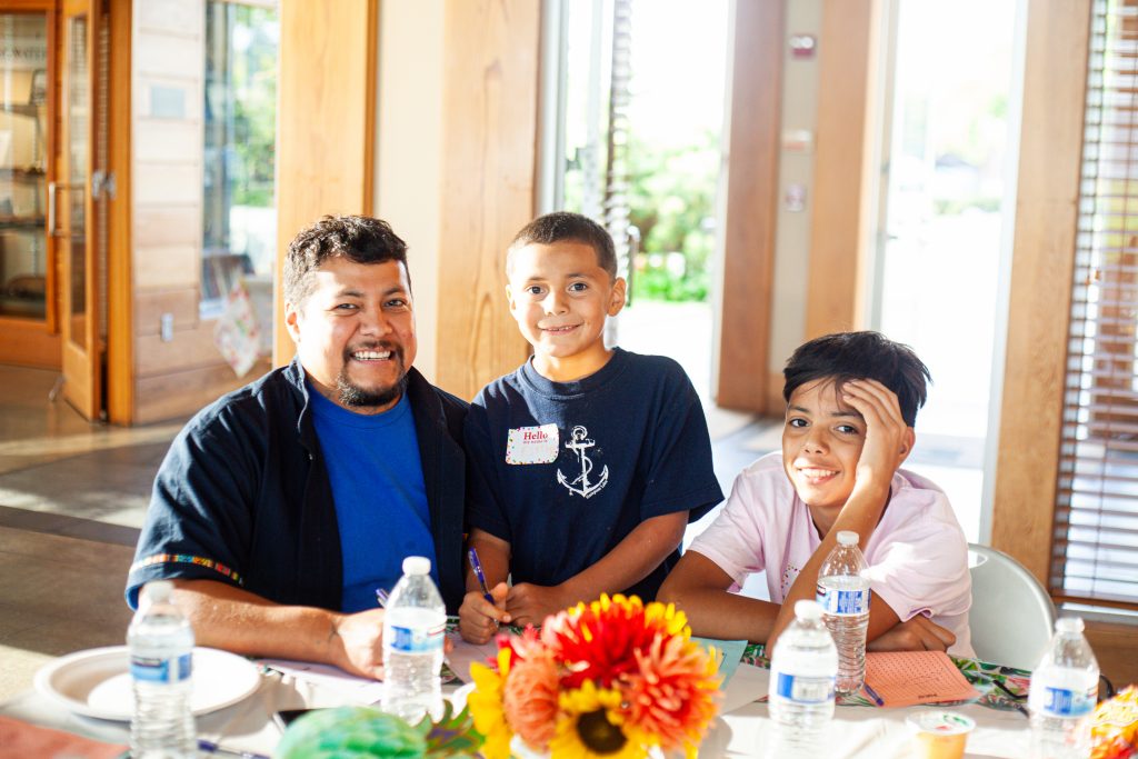 A man and two boys smile as they play games at a table while sun beams in at them from open doors.
