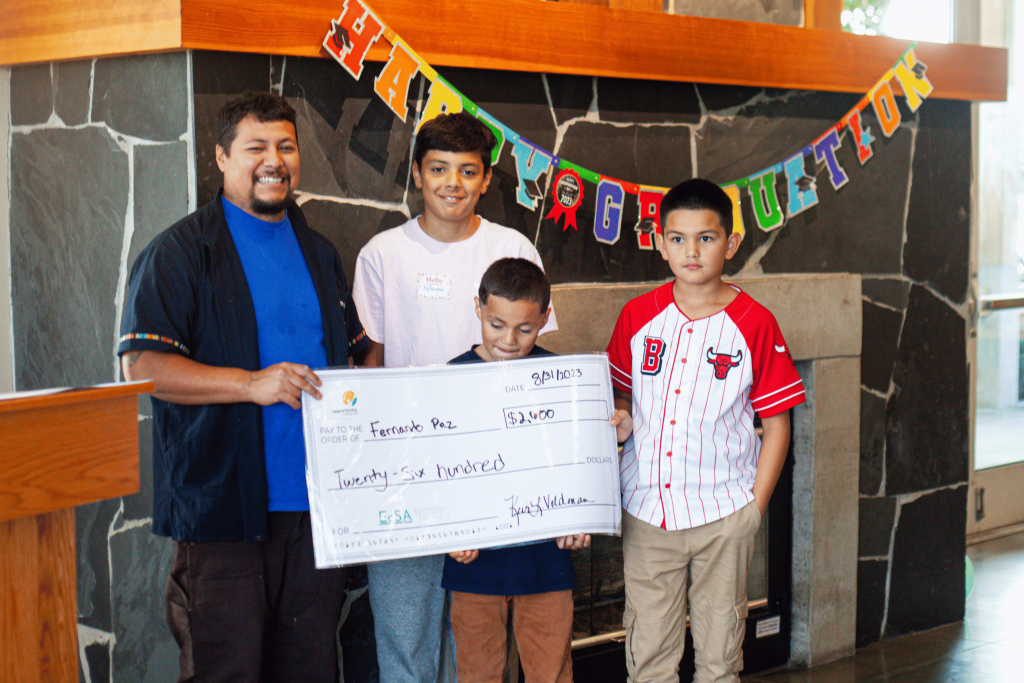 A family of a man and three boys stands with an oversize check that says "8/31/2023: $2,600: Twenty-Six Hundred." In the "For" line, there is the EcSA logo with the acronym in teal. In the top left corner of the check there is the teal and gold Opportunity Council logo.