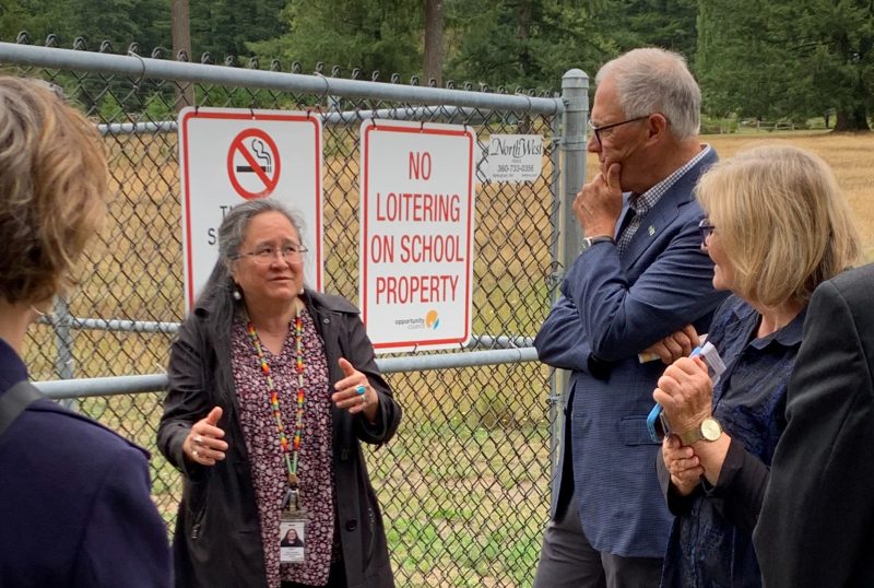 Three people stand around a woman who stands in front of a fence to speak as she gestures to punctuate her words.