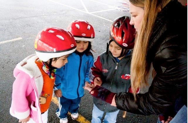 Three young kids in red bike helmets look attentively at something that a woman is showing them in her hand.
