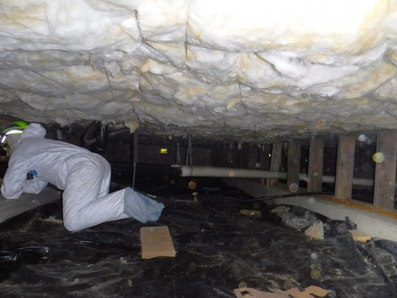 A person wearing a Tyvek suit crawls in a crawl space below a house, which has fiberglass insulation at the top.