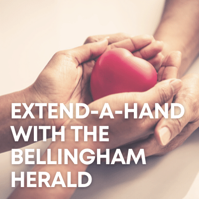 A beige surface has one person's cupped hands in another's, the top hands holding a 3D heart. White text says "Extend-a-Hand with the Bellingham Herald."