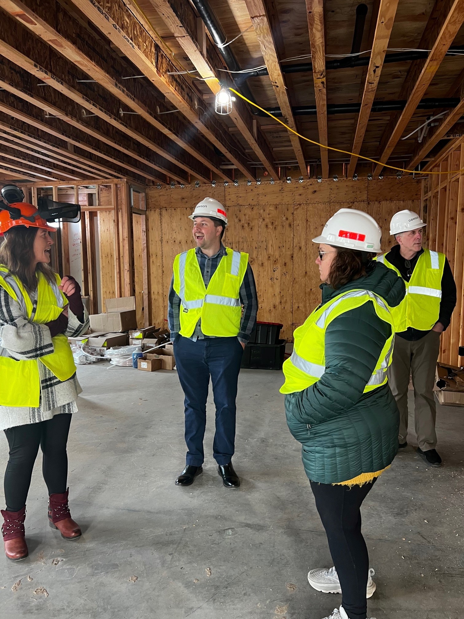 Four people in reflective vests and hard hats stand in a building that is under construction with wood beams on the ceiling and plywood walls.