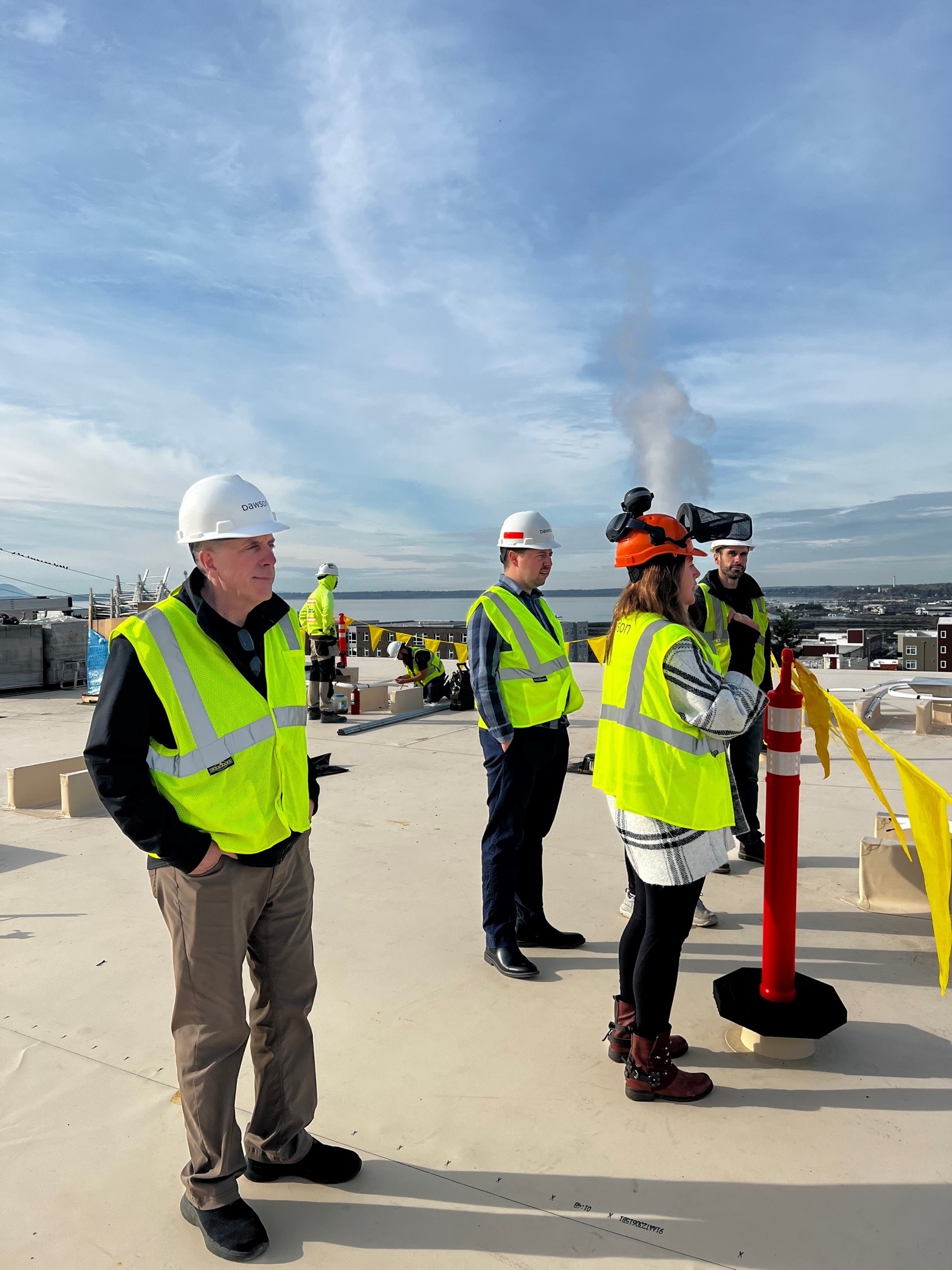 Four people in reflective vests and hard hats stand on the roof of a building that overlooks the Bellingham Bay waterfront.