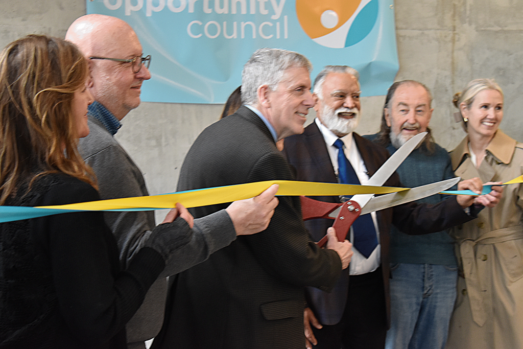 A group of people in formal clothing stand behind teal and yellow ribbon, the man in the center beginning to cut it with giant scissors.