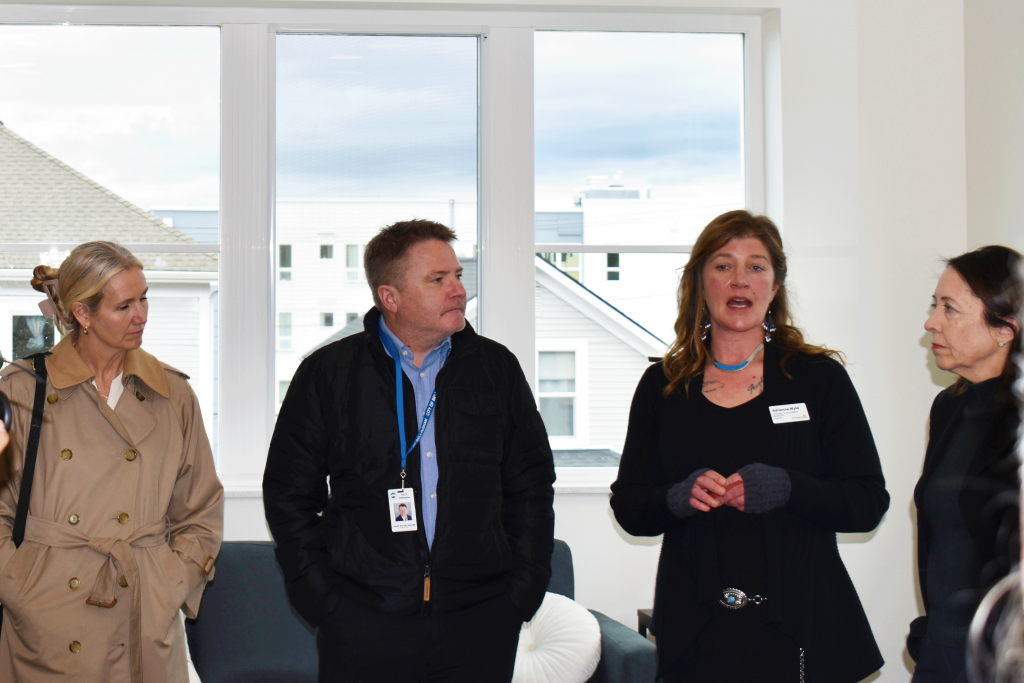 Four people stand together in front of a large bay of windows as one speaks to the rest of the room.