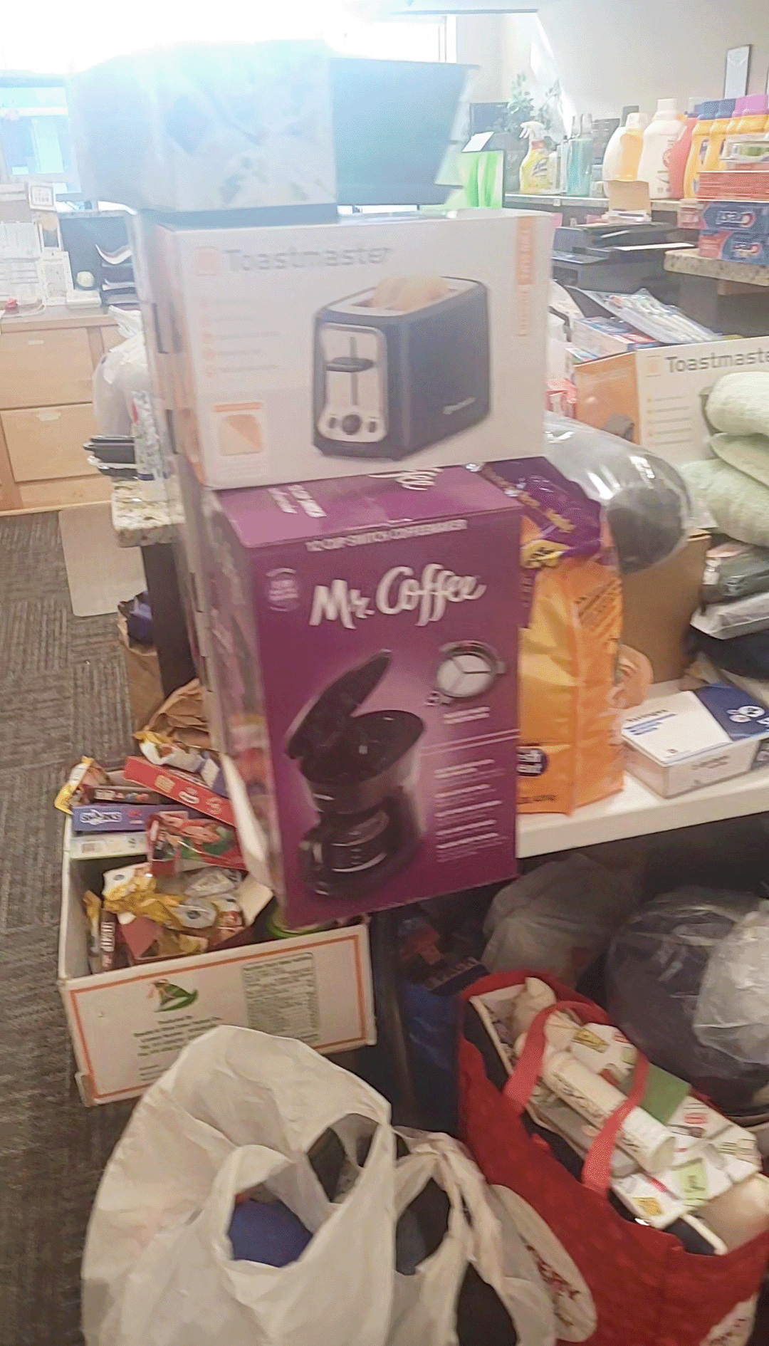 A counter, table, and floor piled high with home items like appliances, food, towels, laundry detergent, and toiletries.