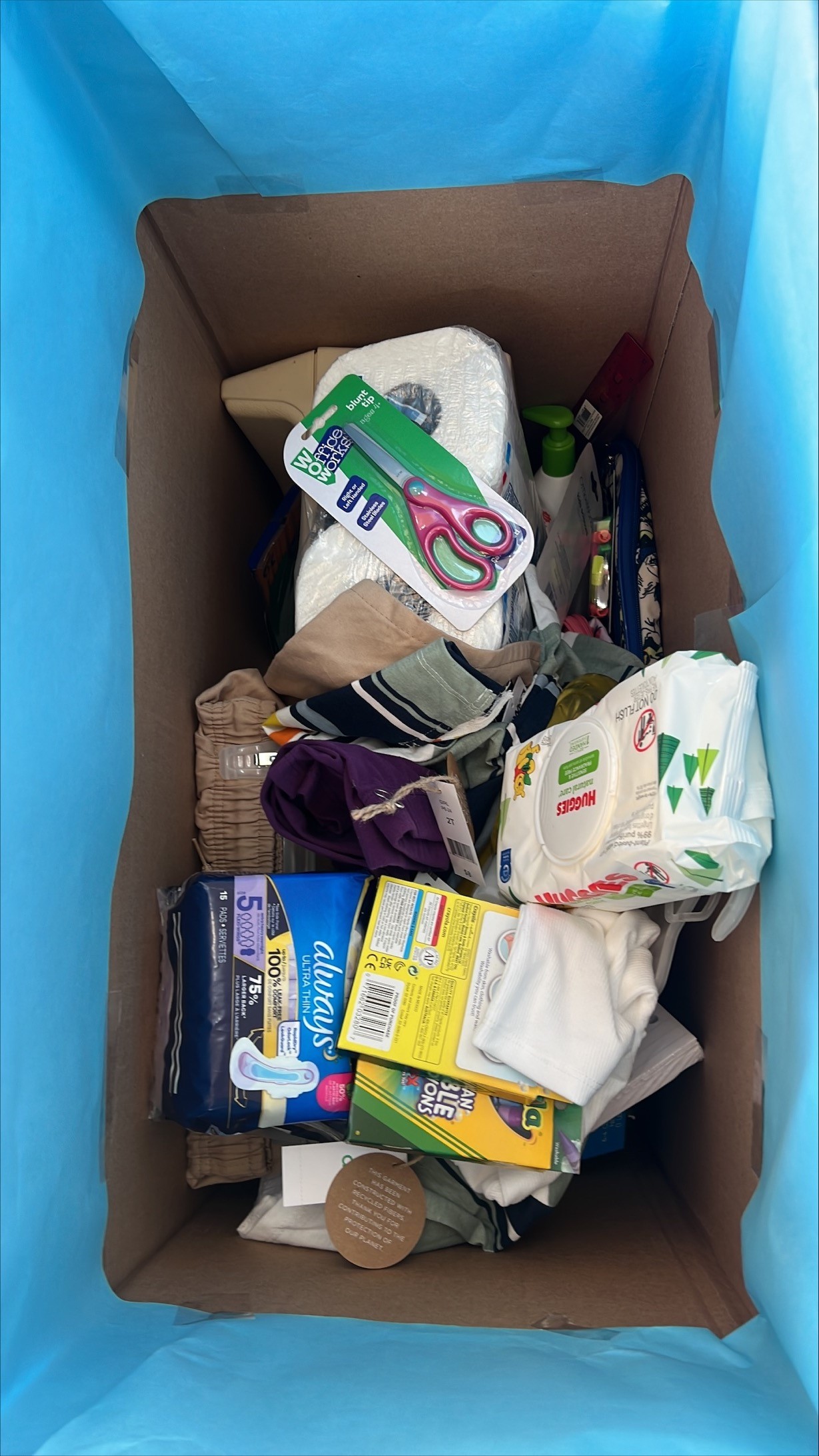 A big box covered in blue paper holds supplies ranging from school supplies to period supplies to baby wipes.