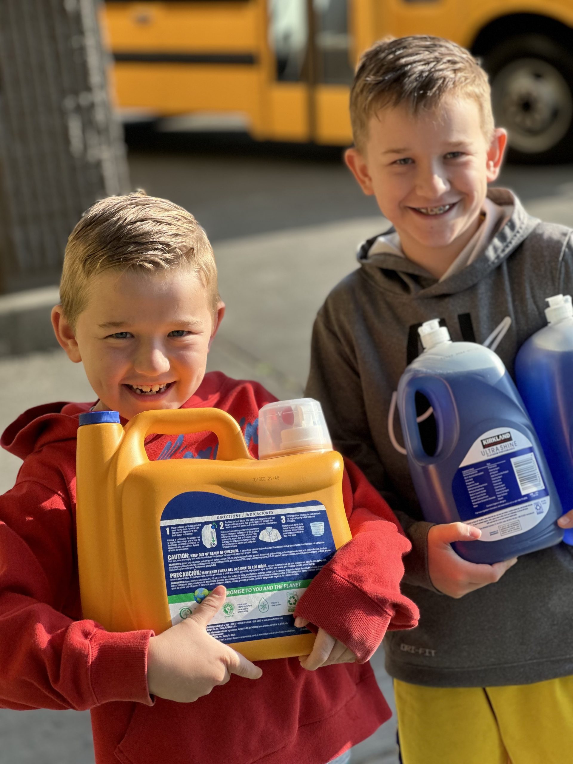 Two kids hold laundry and dish detergent while smiling. A yellow school bus is visible in the far background.