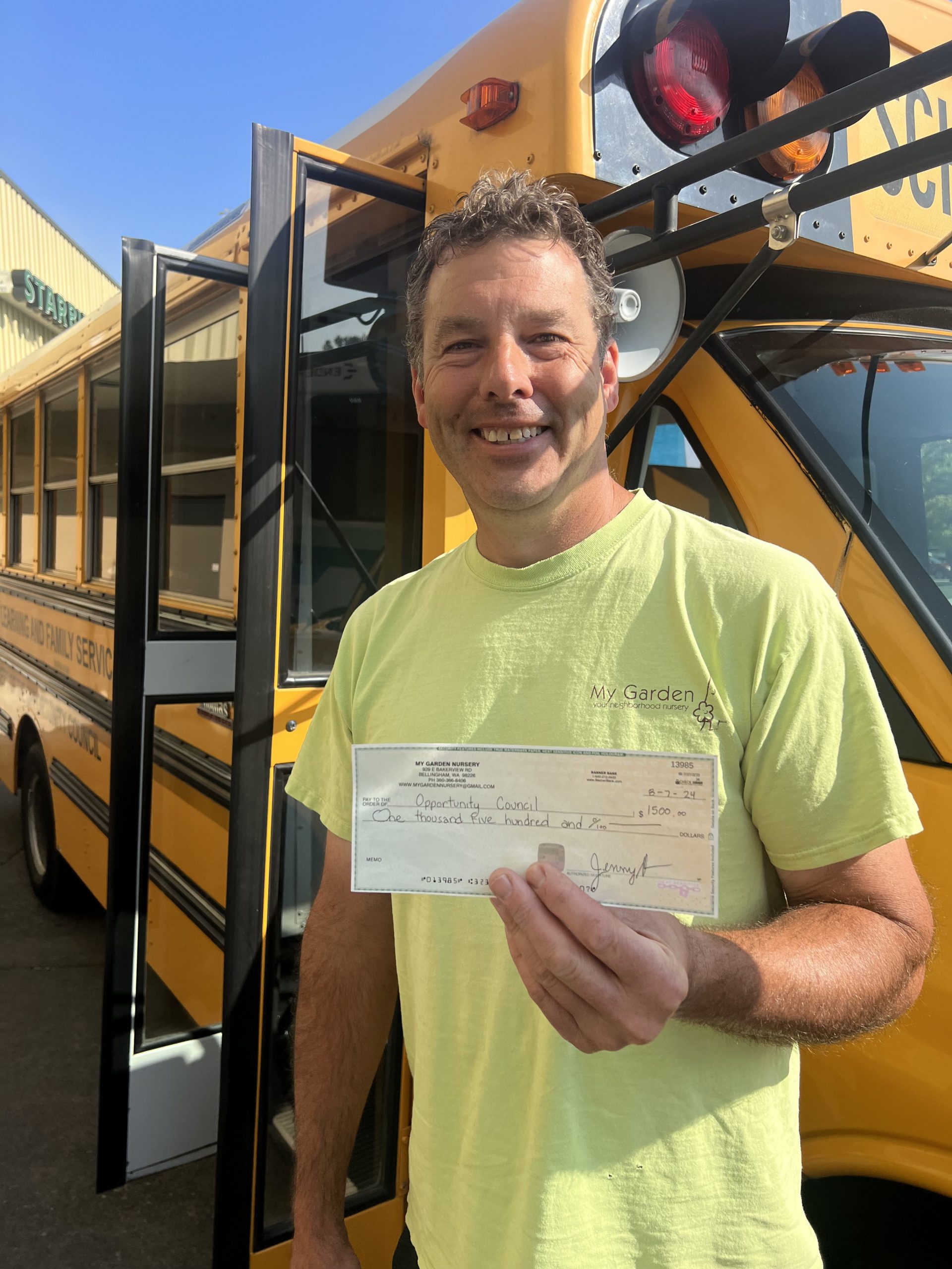 A man stands in front of a yellow school bus, smiling and holding a check.