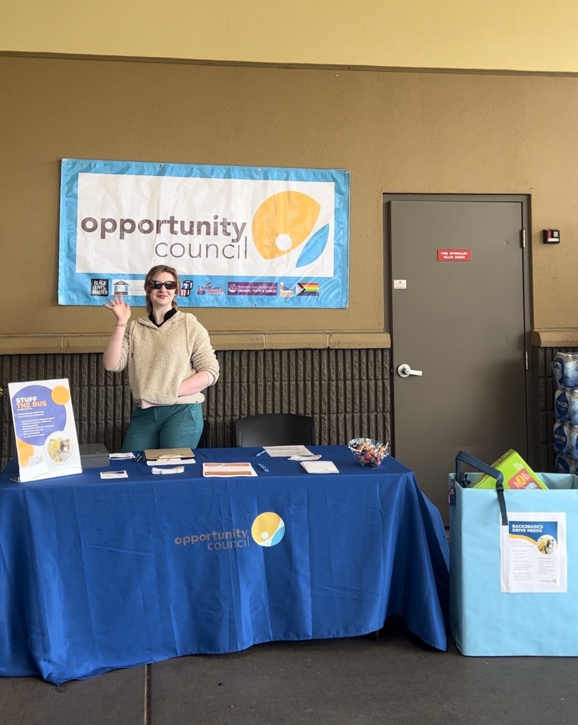 A woman smiles and waves from a table with an Opportunity Council tablecloth that has tabling materials on it and a sign that says "Stuff the Bus." A donation box on the right is filled with stuff. A big banner in the back bears the Opportunity Council logo.