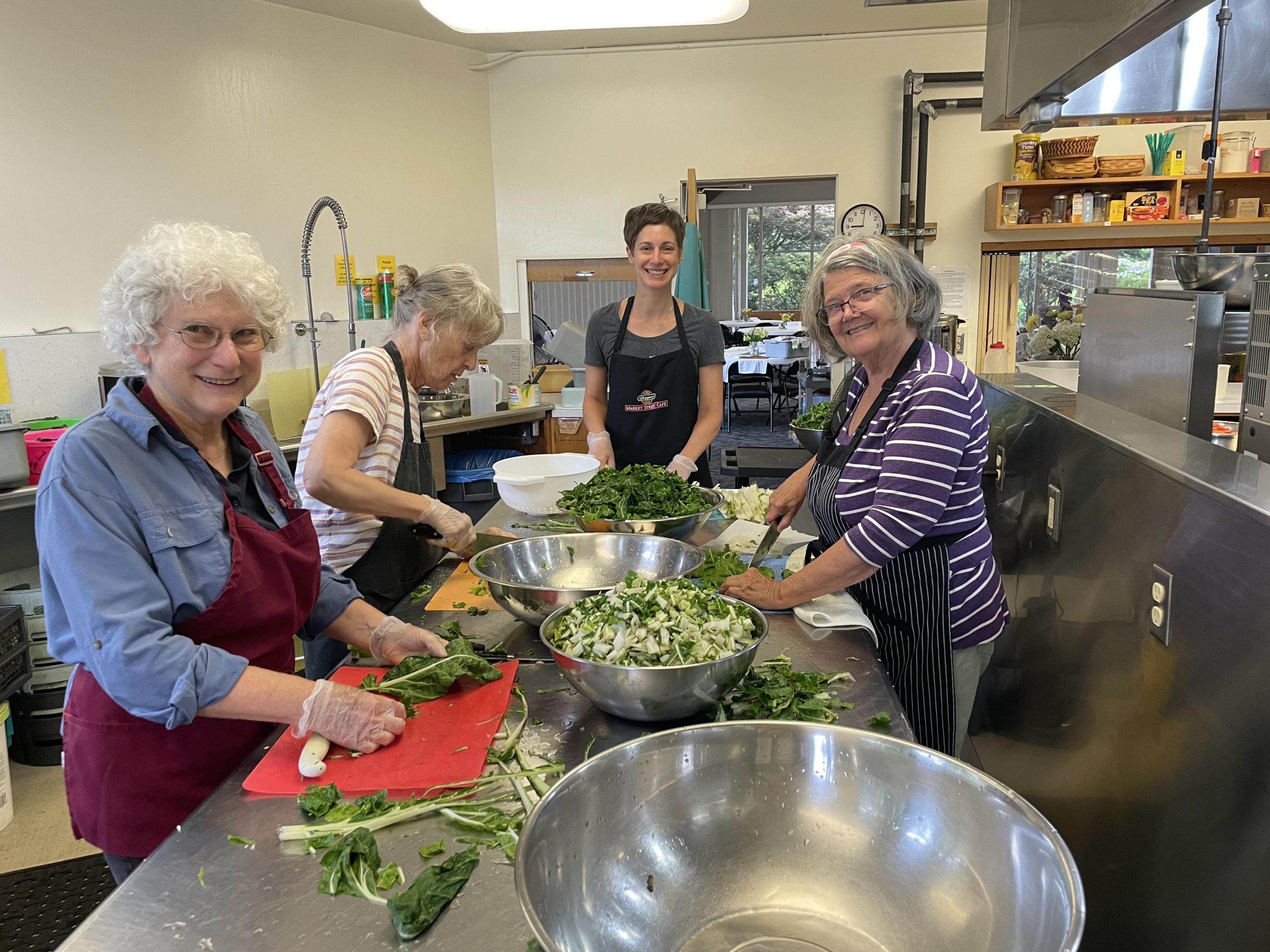 Four people smile around a kitchen prep table while they prepare vegetables.