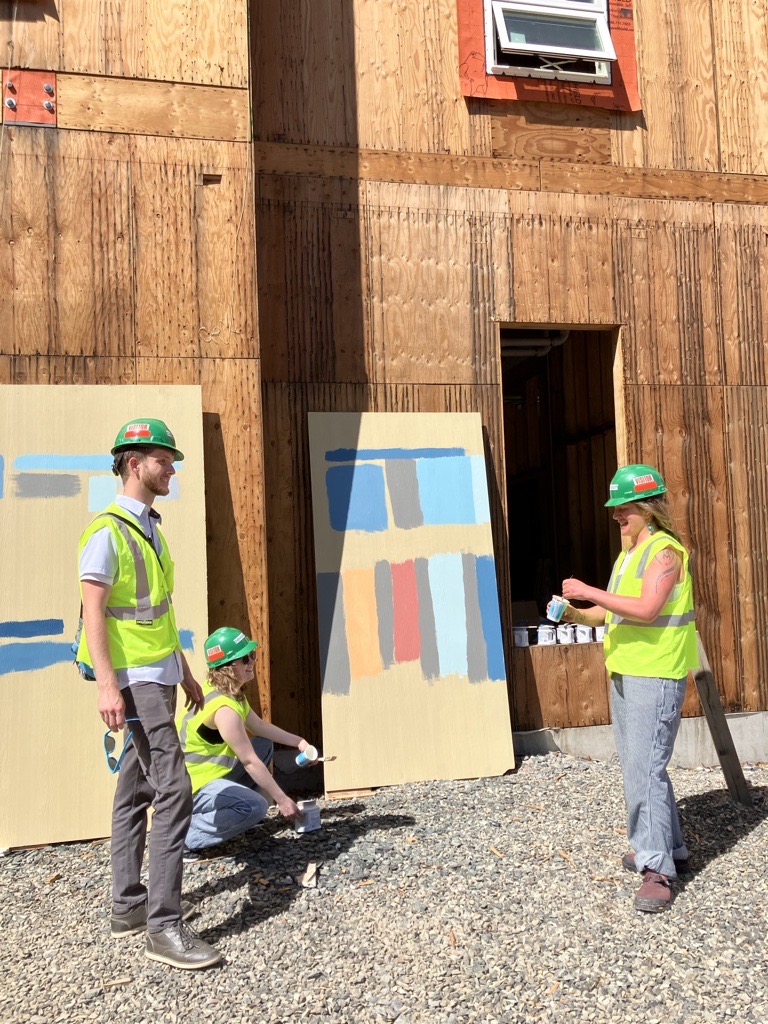 Three people in green hard hats and neon vests stand in front of a piece of plasterboard painted with various swatches, propped up against a building under construction.
