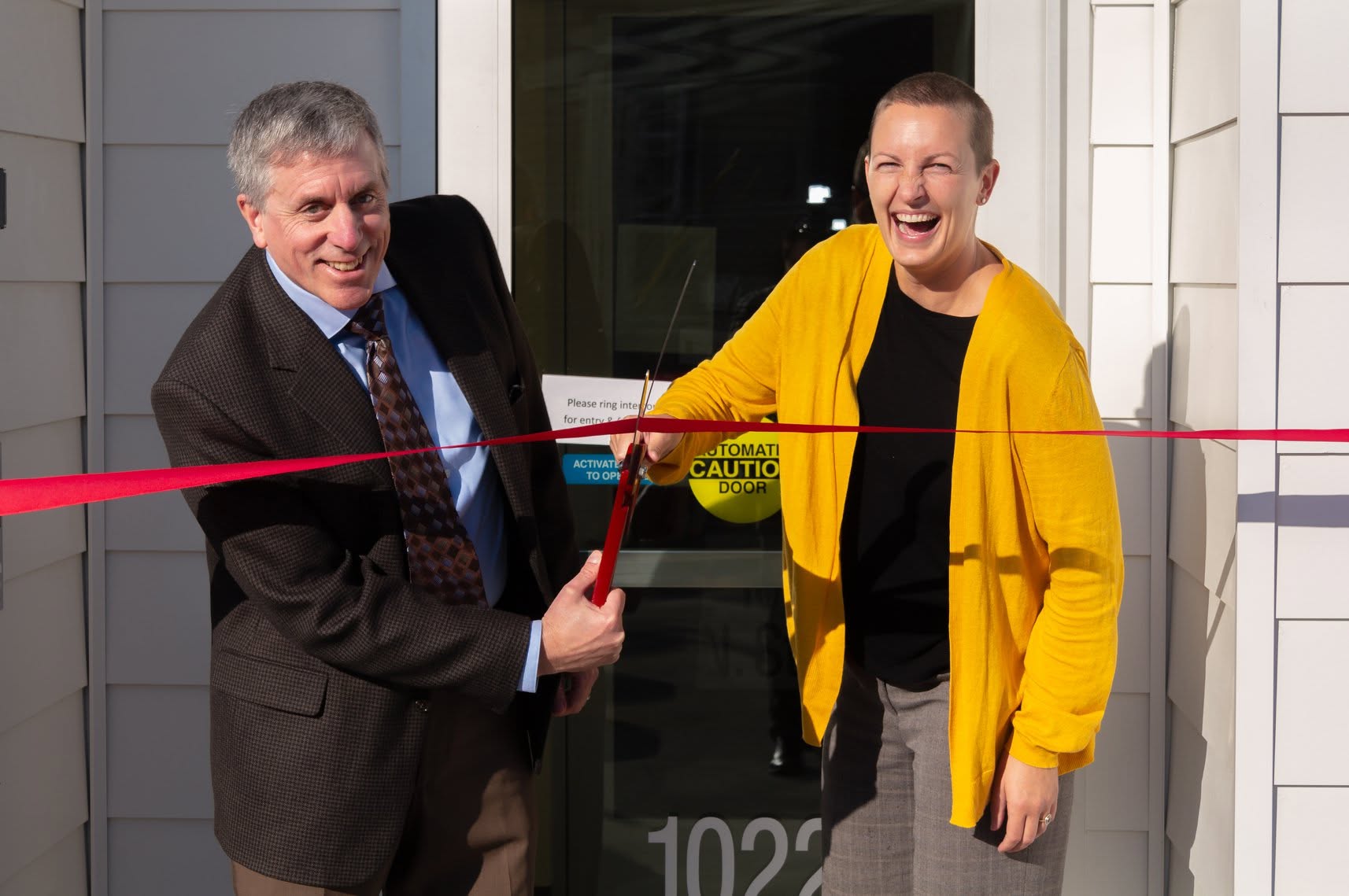 Two people look on joyfully as they cut the ribbon on a new building.