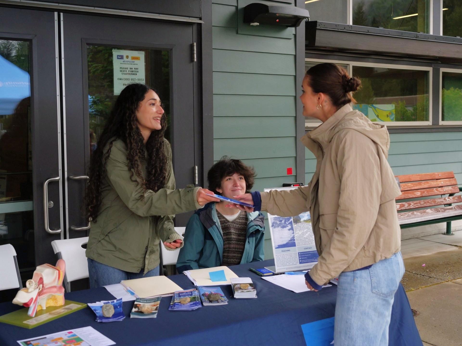 A person behind an info table gives a flyer to a visitor while explaining the info they need to know.