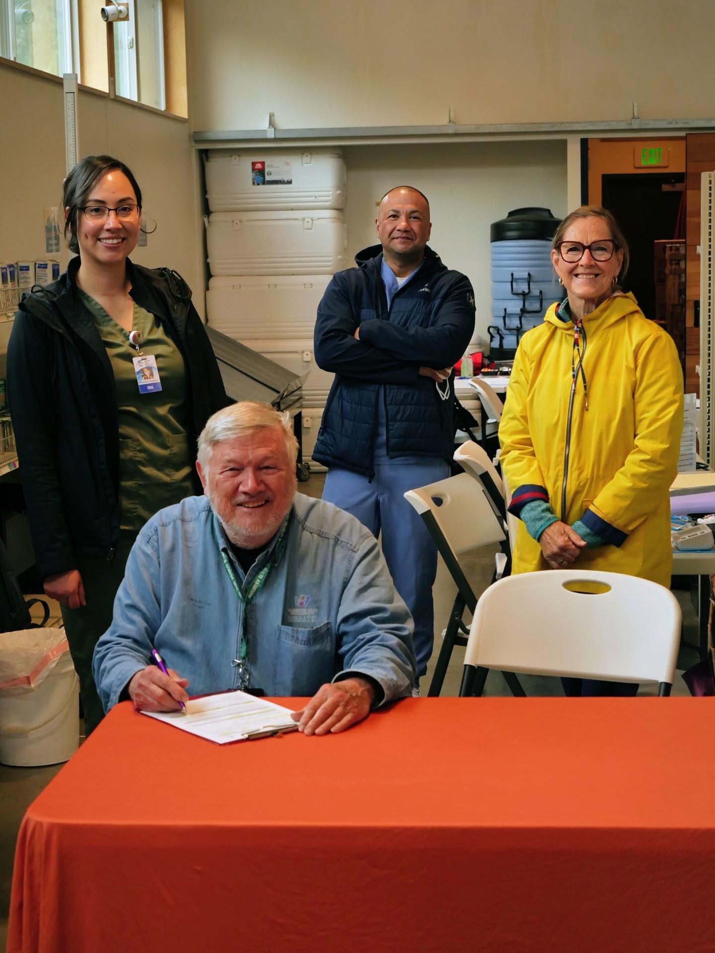 Four people stand or sit behind a table with an orange tablecloth.