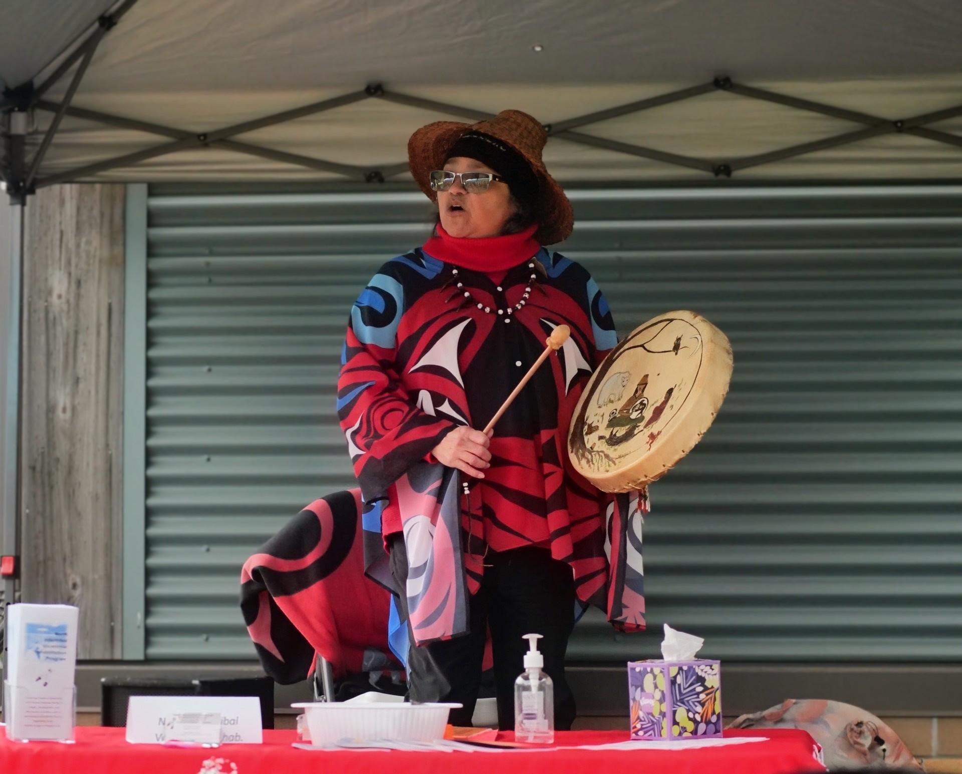 A woman dressed in traditional Native American clothing holds a drum and drumstick