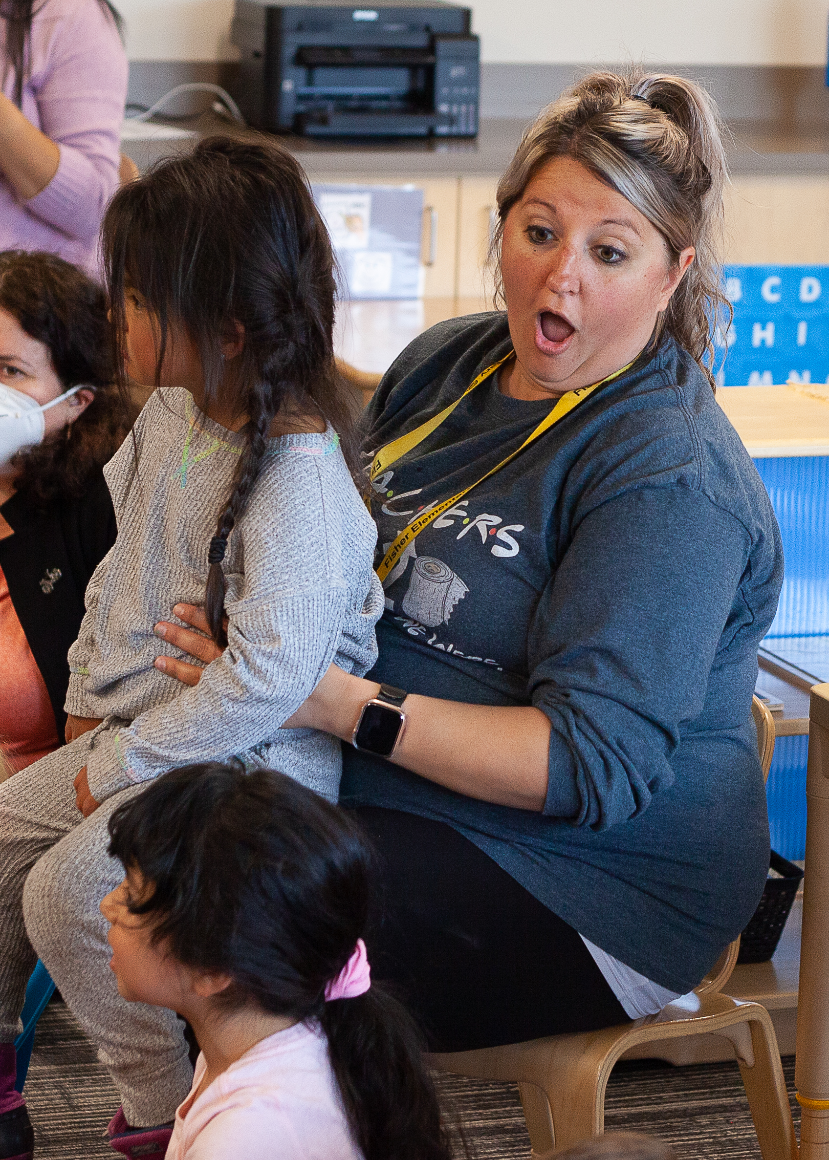 A woman in a classroom with a girl on her lap makes a shocked face